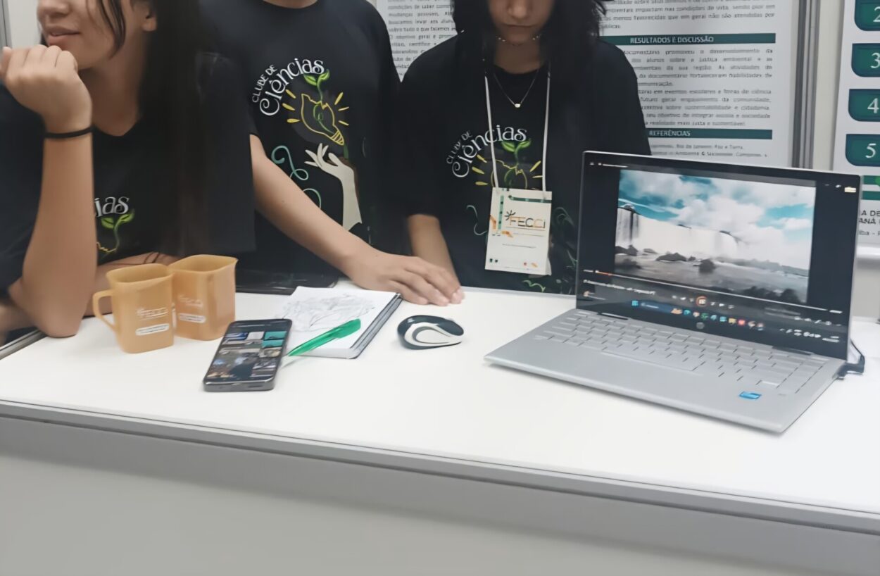 A imagem mostra que um grupo de estudantes está em uma bancada de apresentação, provavelmente em uma feira ou mostra científica. Eles vestem camisetas pretas com a inscrição “Clube de Ciências”. Sobre a mesa há diversos materiais: duas canecas, um celular, um caderno com anotações, uma caneta e um laptop aberto exibindo um vídeo de uma paisagem com água (parece uma cachoeira). Um dos alunos usa um crachá de evento.