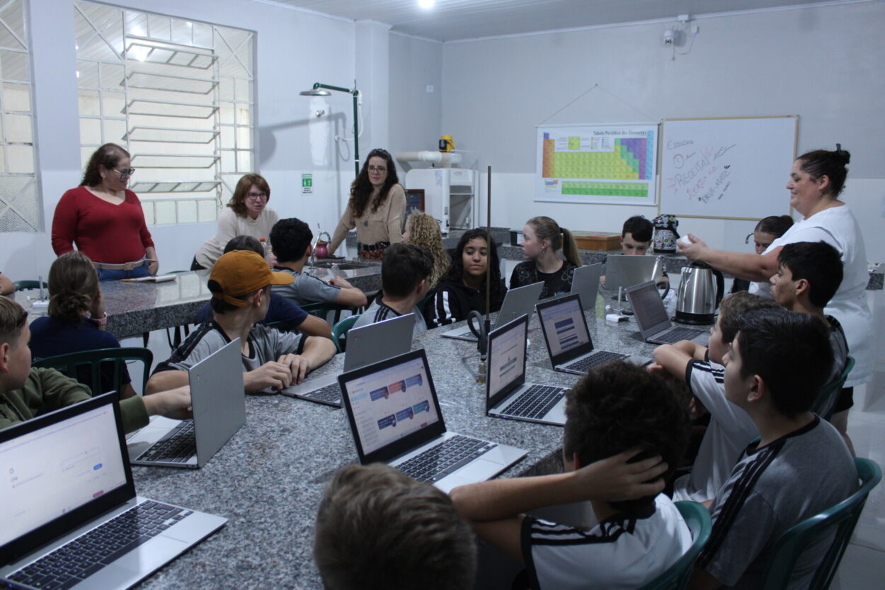 A foto mostra alunos e professora clubista do Clube Ouro Verde, diretora do Colégio Eugênio de Almeida e equipe da UEPG em um laboratório da escola. A sala é branca e bem iluminada, com janelas, quadro, tabela periódica e equipamentos ao fundo. Sentados ao redor de duas bancadas de mármore com vários notebooks, estão os alunos, devidamente uniformizados. Eles observam a diretora falar, ao lado de duas visitantes de pé. A cena ocorreu durante um discurso da diretora sobre a importância do trabalho dos clubistas, que na foto parecem atentos à fala.
