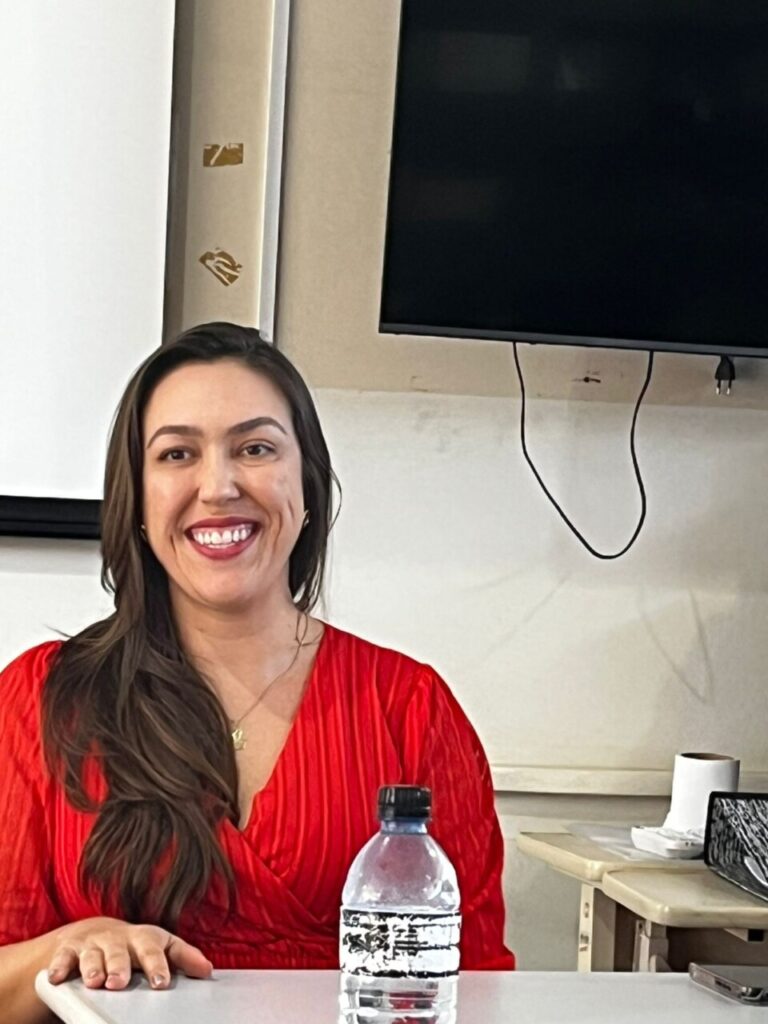 A imagem mostra uma mulher sorrindo, sentada em um ambiente interno que parece ser uma sala de reunião ou escritório. Ela tem cabelos longos e escuros, usa uma blusa vermelha de tecido brilhante e um colar delicado. À frente dela, sobre a mesa, aparece parcialmente uma garrafa plástica transparente. No fundo da imagem há uma parede clara, uma televisão ou monitor preso na parede com fios aparentes e, à direita, uma pequena mesa com uma xícara branca e outros objetos. O clima da foto é descontraído e acolhedor.