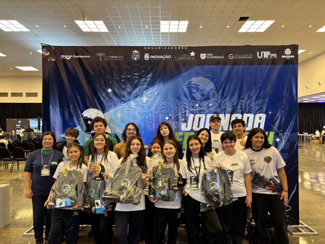 Foto de um grupo de cerca de 15 pessoas, entre crianças, adolescentes e adultos, reunido em frente a um grande painel do evento Jornada Educatech. Alguns participantes seguram troféus e kits de premiação embalados em plástico. O grupo está sorrindo e posando para a foto. Ao fundo, o painel azul e preto traz elementos relacionados à tecnologia e inovação, além dos logotipos dos organizadores. A foto foi feita em um amplo espaço interno, com outras pessoas e estandes do evento ao fundo.