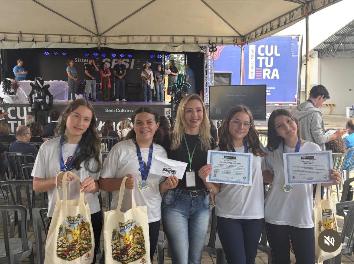 Foto tirada em uma feira científica, em um espaço coberto com palco ao fundo. Na frente, cinco jovens e uma mulher adulta posam lado a lado, sorrindo para a câmera. As jovens usam camisetas brancas e algumas têm medalhas no pescoço; duas seguram certificados e outras carregam sacolas de tecido com ilustrações coloridas. A mulher ao centro usa blusa preta, calça jeans e um crachá. Ao fundo, há um palco com várias pessoas em pé, iluminação azul e um painel com a palavra “Cultura”. O ambiente indica uma premiação ou apresentação dentro da feira científica.