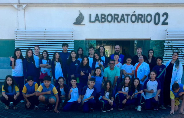 Foto de grupo tirada em frente ao prédio identificado pela placa “Laboratório 02”. Cerca de 30 pessoas, entre adolescentes e adultos, posam reunidas ao ar livre sobre um piso de blocos intertravados. A maioria veste uniformes escolares em tons de azul, branco e amarelo, enquanto alguns adultos usam jalecos ou roupas casuais. O grupo está distribuído em pé e agachado, formando várias fileiras. Algumas pessoas sorriem e fazem gestos com as mãos para a câmera. Ao fundo, a fachada clara do laboratório possui detalhes em verde e estruturas metálicas brancas nas laterais. A iluminação sugere um dia ensolarado, com sombras suaves e tonalidade azulada na imagem.