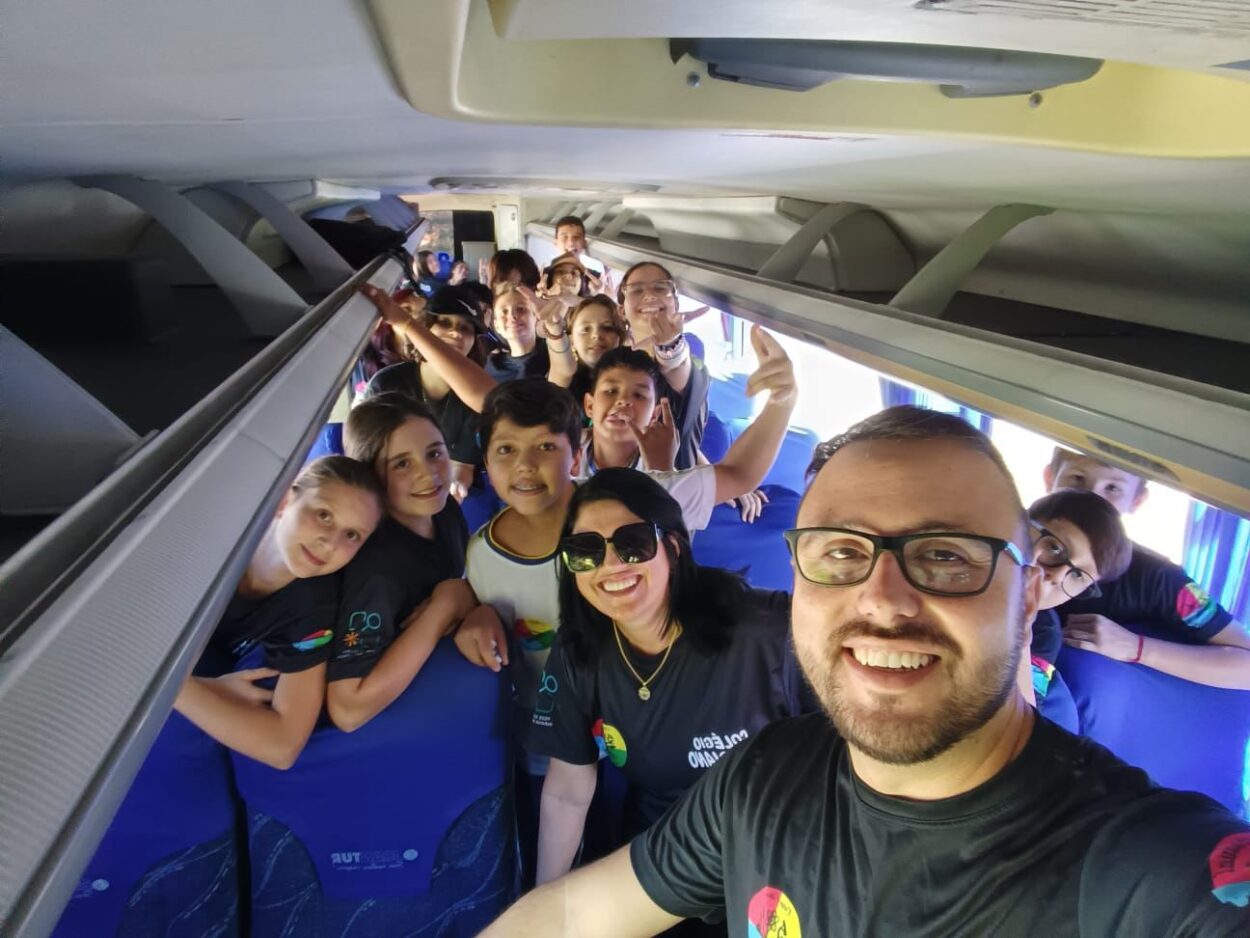 Foto dentro de um ônibus durante uma viagem escolar. Em primeiro plano, um homem de óculos, barba curta e camiseta preta faz uma selfie sorrindo. Ao lado dele, uma mulher de óculos escuros também sorri para a câmera. Ao fundo, várias crianças aparecem sentadas e em pé entre os bancos do ônibus, algumas sorrindo, fazendo sinais com as mãos e olhando para a foto. Todos usam camisetas pretas semelhantes, indicando participação em um mesmo grupo ou atividade. O clima é descontraído e alegre