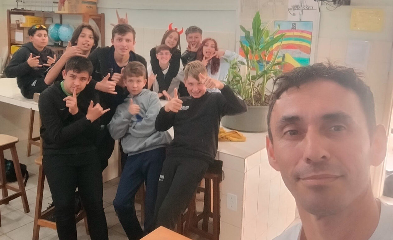 Foto em grupo tirada em ambiente escolar ou laboratório, com um professor em primeiro plano fazendo uma selfie e vários adolescentes ao fundo. O grupo posa de forma descontraída e divertida, fazendo sinais com as mãos, mostrando joinhas e expressões brincalhonas. O ambiente possui bancadas brancas, banquetas de madeira, prateleiras com materiais e globos terrestres. Ao lado direito há vasos com plantas e um cartaz colorido com um arco-íris desenhado. A iluminação é suave e quente, transmitindo clima de amizade, interação e descontração entre a turma.