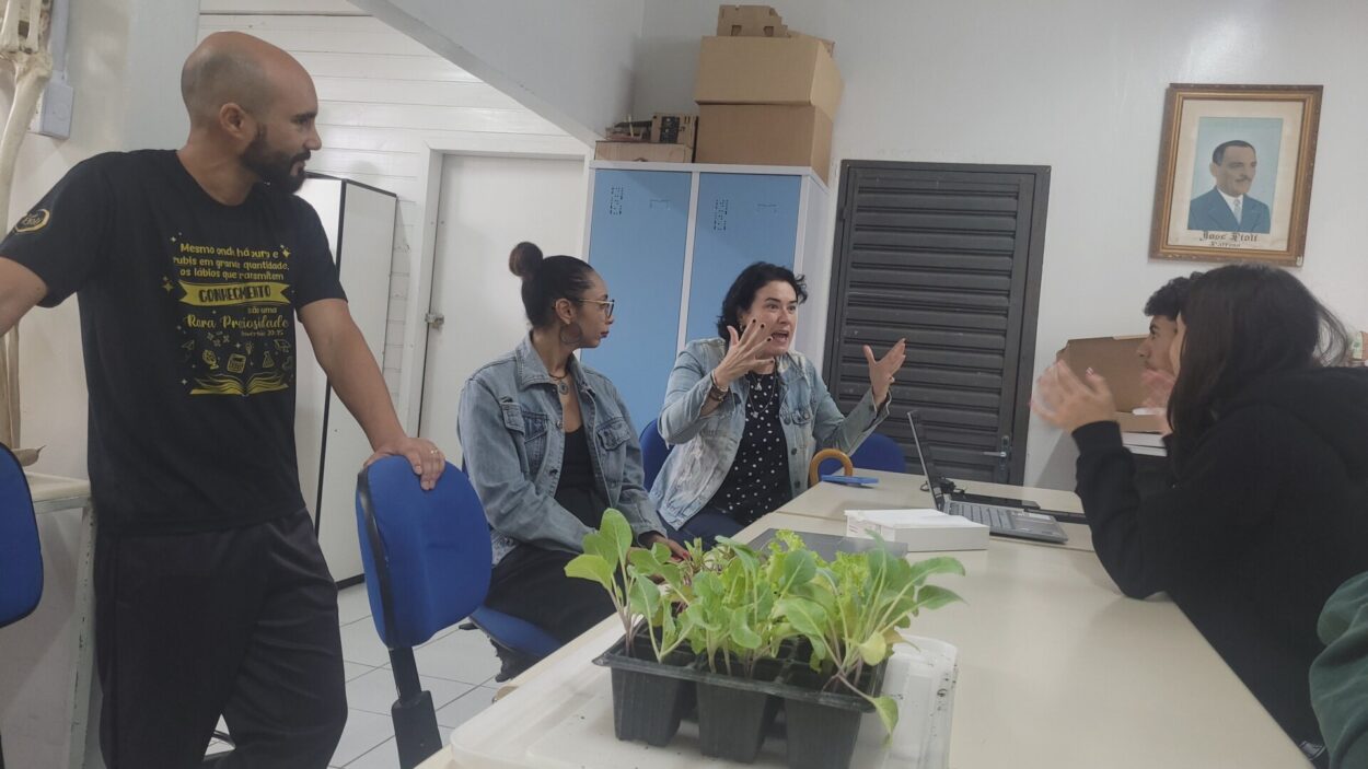 Em uma sala de aula, quatro pessoas conversam ao redor de uma mesa enquanto estudantes acompanham a atividade. Sobre a mesa há bandejas com mudas de plantas, papéis, um notebook e materiais de apoio. Ao fundo aparecem armários, caixas e um quadro na parede.