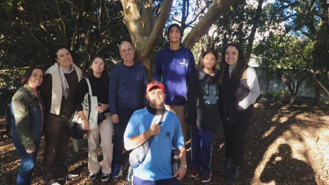 Oito pessoas posam para uma foto em uma área com árvores e vegetação. O grupo está reunido em frente a uma árvore grande, em um ambiente externo com chão coberto por folhas secas. Algumas pessoas estão em pé e uma está ajoelhada à frente do grupo.