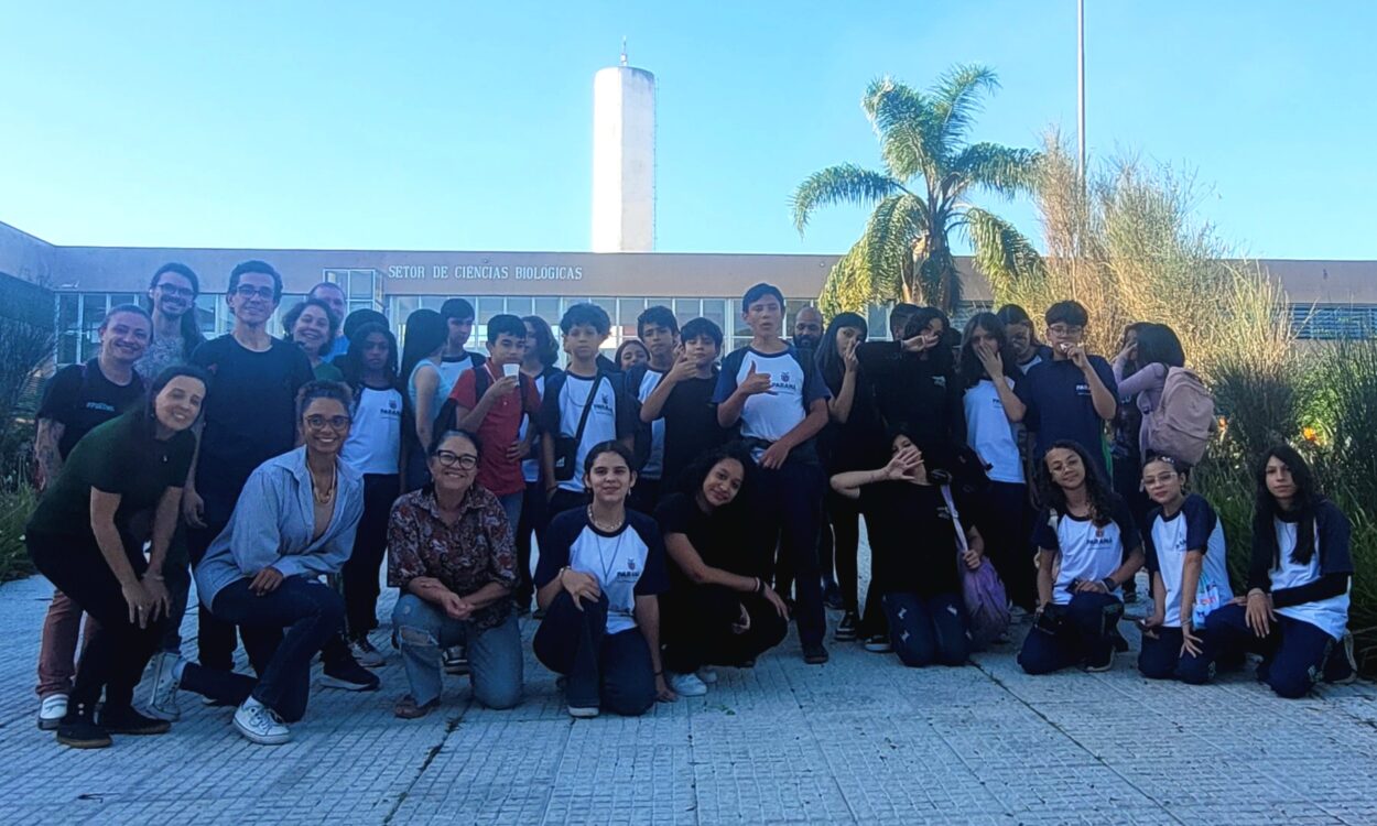 Grupo grande de estudantes e professores posando juntos ao ar livre, em frente a um prédio com a inscrição “Setor de Ciências Biológicas”. As pessoas estão organizadas em duas fileiras, algumas em pé e outras agachadas na frente, sorrindo para a câmera. A maioria veste roupas casuais, com várias camisetas escolares brancas com detalhes escuros. Ao fundo, há uma palmeira, vegetação e uma estrutura alta, sob um céu azul claro, indicando um dia ensolarado.