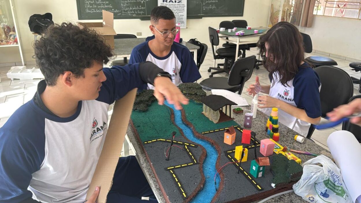 Fotografia em plano médio de três estudantes (dois rapazes e uma moça) trabalhando juntos ao redor de uma mesa de granito em uma sala de aula ou laboratório escolar. Eles usam uniformes escolares da rede pública do Paraná, que consistem em camisetas brancas com mangas azuis e o brasão do estado no peito. Sobre a mesa, há uma maquete detalhada construída sobre uma base retangular escura, retratando o fluxo e a aplicação de um sistema de biodigestor integrado a uma comunidade rural e urbana. A maquete apresenta os seguintes elementos: O biodigestor e a área rural: No quadrante superior direito da maquete, há uma estrutura miniatura coberta por um telhado escuro curvado, simulando o galpão de produção (como uma granja) conectado ao sistema de biodigestor para captação de resíduos orgânicos. Próximo a ele, áreas verdes de textura rugosa representam a vegetação ou plantações que se beneficiam do biofertilizante. O curso de água: Uma linha azul texturizada cruza a maquete diagonalmente, simulando um rio ou riacho que corta o terreno, dividindo a área de produção da zona urbana. A zona urbana e caminhos: Na parte inferior e à direita, há pequenas casas coloridas feitas de papel cartolina (em tons de laranja, amarelo, rosa e verde), ruas de asfalto preto com linhas tracejadas amarelas e pequenos postes de luz com fios expostos conectados, ilustrando a distribuição da energia elétrica ou biogás gerados a partir do biodigestor para a comunidade.
