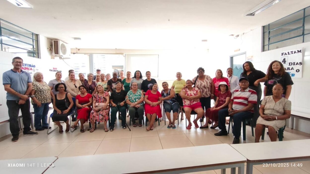 Fotografia em ambiente interno mostra um grupo de cerca de 30 pessoas reunidas em uma sala ampla e iluminada. A maioria são mulheres idosas, acompanhadas por alguns homens e pessoas mais jovens. Parte do grupo está sentada em cadeiras organizadas em semicírculo, enquanto outras pessoas permanecem em pé ao fundo. As participantes usam roupas coloridas e casuais, com vestidos estampados, blusas vermelhas, verdes e pretas. Algumas sorriem para a câmera. O clima da imagem transmite acolhimento, convivência e encontro comunitário. O espaço possui paredes claras, janelas grandes nas laterais e cartazes afixados. À direita, um cartaz escrito “FALTAM 4 DIAS PARA O ENEB” aparece na parede. Na frente da foto há mesas brancas vazias. No canto inferior esquerdo aparece a marca d’água da câmera do celular, e no canto inferior direito a data e horário do registro: 23 de outubro de 2025, às 14h49.