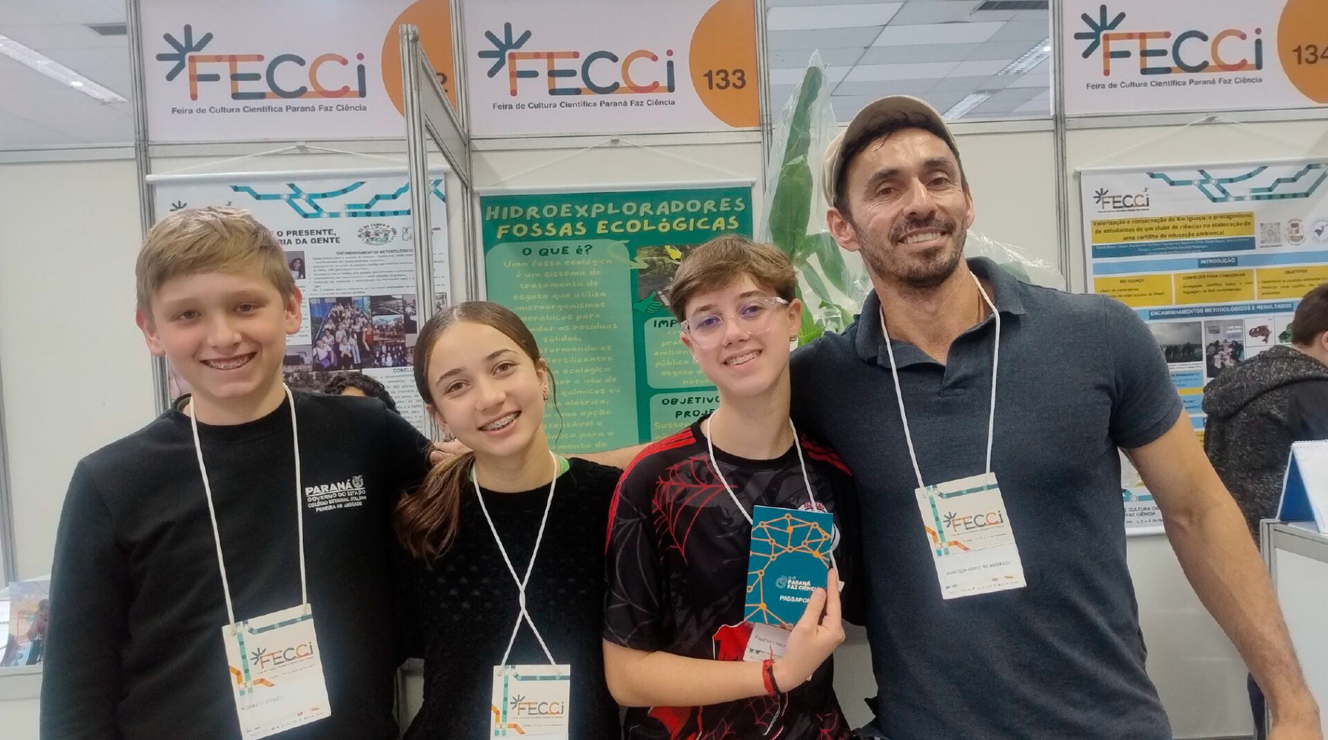 Quatro pessoas posam sorrindo em um estande da FECCI (Feira de Cultura Científica Paraná Faz Ciência). Três adolescentes e um professor estão lado a lado, usando crachás de identificação pendurados no pescoço. Ao fundo, há painéis com informações e banners de projetos científicos, incluindo um cartaz verde com o título “Hidroexploradores Fossas Ecológicas”. O adolescente à esquerda usa camiseta preta com o logotipo do Governo do Paraná. A menina ao centro veste blusa preta e tem o cabelo preso. A adolescente ao lado dela usa óculos, camiseta preta com detalhes vermelhos e segura um livreto azul. O homem à direita veste camisa polo escura e boné claro. Todos demonstram expressão alegre e orgulhosa, em um ambiente de exposição científica escolar.