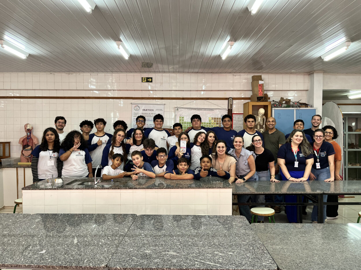 Uma turma grande de estudantes e professores posa para uma foto dentro de um laboratório escolar. Eles estão alinhados atrás de uma bancada comprida de pedra, com alguns alunos apoiados nela na frente. A maioria usa camisetas claras com mangas escuras, de uniforme. O ambiente tem paredes revestidas de azulejos brancos e várias luzes fluorescentes no teto. Ao fundo, há materiais de estudo, como um esqueleto humano em pé e um modelo do corpo humano, além de armários e cartazes. Todos estão próximos, muitos sorrindo e olhando para a câmera, transmitindo um clima de grupo unido e descontraído.