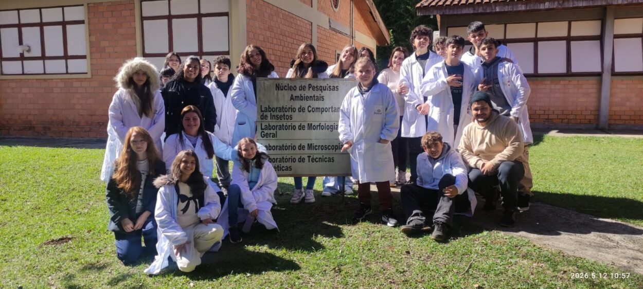 Fotografia tirada ao ar livre, em frente a um prédio de laboratórios com paredes de tijolos aparentes. Um grupo de aproximadamente vinte estudantes e professores posa para a foto sobre um gramado, a maioria vestindo jalecos brancos. No centro, os participantes seguram uma placa com os dizeres: “Núcleo de Pesquisas Ambientais”, seguida dos nomes de laboratórios ligados à área ambiental e biológica. O grupo sorri para a câmera em um dia ensolarado, durante uma atividade de visita ou prática acadêmica. Ao fundo, aparecem as edificações do campus e áreas verdes.