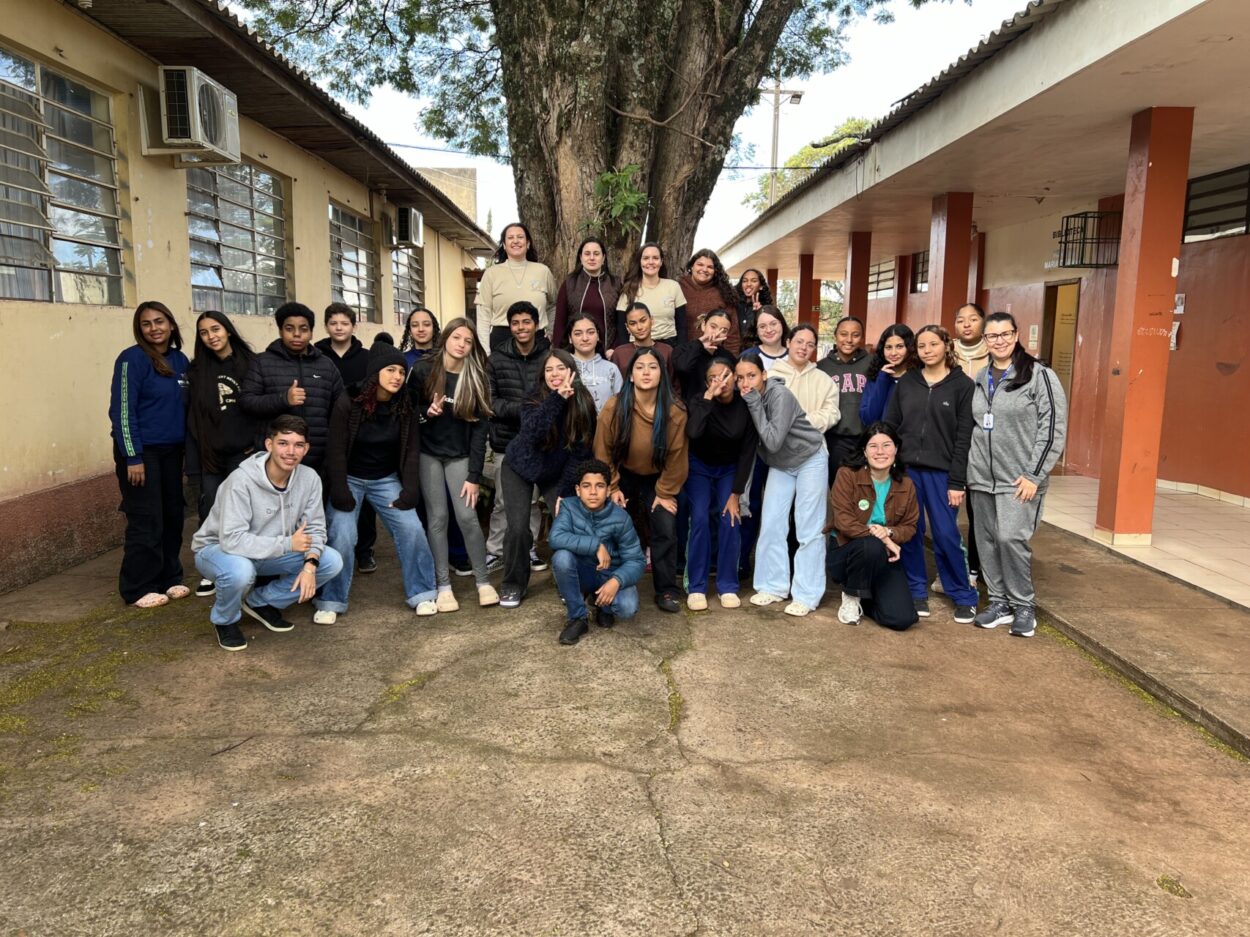 A imagem mostra uma foto de um grupo ao ar livre, registrada em um pátio escolar. Ao centro da cena há uma grande árvore de tronco largo, que domina o fundo da fotografia e serve como ponto de referência para o grupo. Ao todo, há cerca de 30 pessoas, entre adolescentes e alguns adultos, organizadas em três fileiras. A maioria dos estudantes está em pé, enquanto alguns estão agachados ou ajoelhados na parte da frente.
Os estudantes vestem roupas casuais e de inverno, como casacos, moletons e calças jeans ou de uniforme escolar. Alguns usam gorros e crocs. A iluminação é natural, indicando um dia claro, com sombras suaves projetadas pela árvore e pelos prédios.