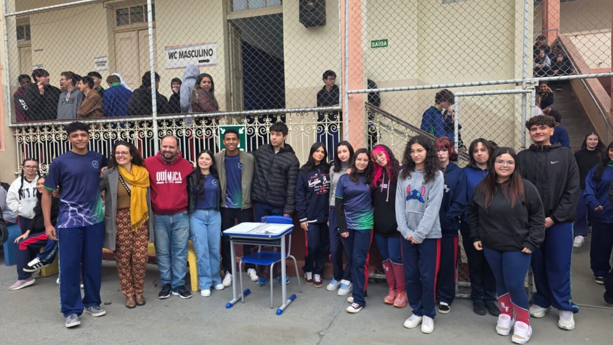 A foto mostra um grupo de aproximadamente vinte estudantes e três adultos posando para uma fotografia no pátio de uma escola. A maioria está em pé, lado a lado, olhando para a câmera. Os estudantes usam uniformes em tons de azul, enquanto os adultos vestem roupas casuais. Ao centro da imagem há uma carteira escolar azul com alguns documentos sobre ela. Ao fundo, aparecem outros alunos circulando pelos corredores e escadas da escola, delimitados por grades metálicas. O ambiente é aberto, iluminado pela luz do dia, e transmite um clima de confraternização e celebração.