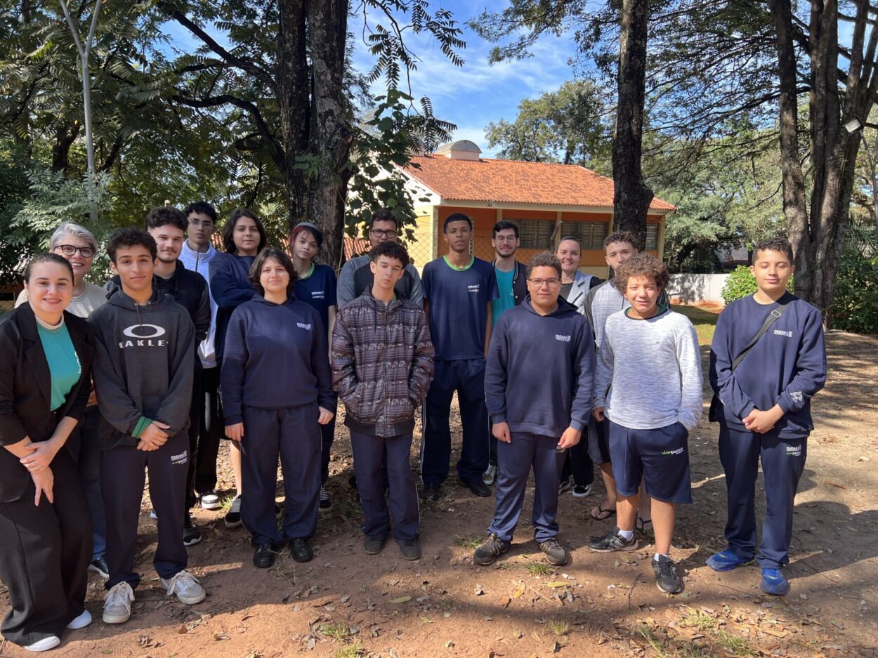 A imagem mostra um grupo de aproximadamente 15 pessoas, entre estudantes e professores, posando para uma foto ao ar livre em um ambiente escolar. Os participantes estão organizados em duas fileiras, olhando para a câmera. A maioria dos estudantes veste uniforme esportivo azul-marinho, enquanto alguns usam moletons e casacos. À esquerda da imagem, há duas mulheres adultas sorrindo. O cenário é cercado por árvores, criando um ambiente arborizado e agradável. A luz do sol ilumina o grupo, projetando sombras suaves no chão de terra. A fotografia transmite um clima de integração, participação e convivência escolar, sugerindo um momento de atividade coletiva.