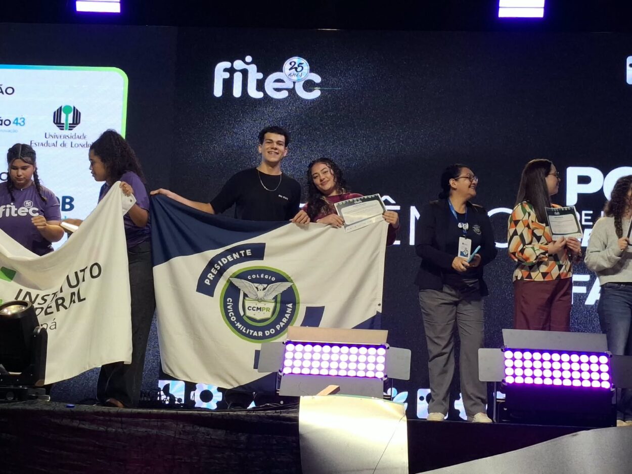 Fotografia da cerimônia de premiação da FITEC. Sobre o palco, estudantes e representantes de instituições de ensino seguram certificados e bandeiras de suas escolas. Ao centro, dois estudantes exibem a bandeira do Colégio Cívico-Militar Presidente Kennedy, de Rolândia, enquanto uma das estudantes segura um certificado. Ao fundo, um painel escuro apresenta o logotipo da FITEC, e os refletores iluminam a parte da frente do palco.
