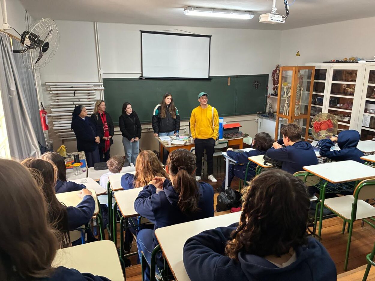 Fotografia de uma sala de aula. Os estudantes estão sentados em carteiras, voltados para a frente da sala, onde cinco pessoas estão em pé realizando uma apresentação. Ao centro, um jovem de moletom amarelo fala para a turma, acompanhado por quatro mulheres ao seu lado. Ao fundo, há um quadro verde, uma tela de projeção recolhida e armários com materiais didáticos e modelos anatômicos. A sala é iluminada por luzes fluorescentes no teto, e os alunos acompanham atentamente a atividade.