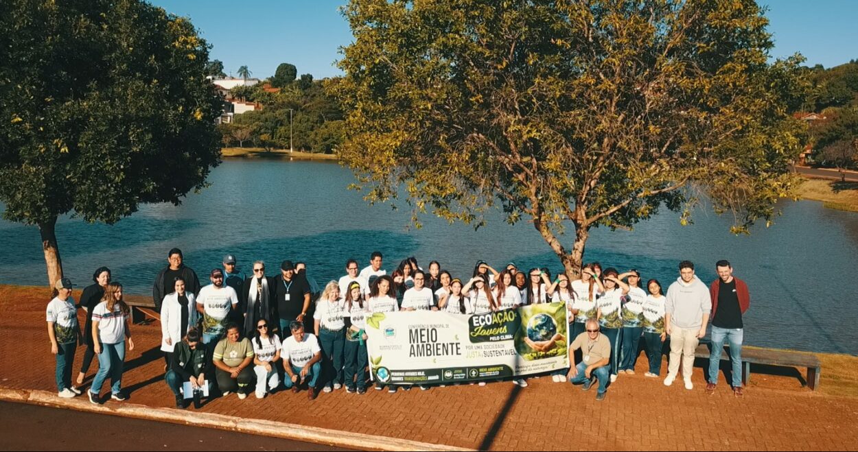 Uma fotografia ao ar livre mostra um grupo de cerca de quarenta pessoas, em sua maioria jovens e adolescentes, reunidas para uma foto coletiva à margem de um grande lago. O cenário ao fundo exibe uma vasta extensão de água calma sob um céu azul, com árvores frondosas e casas na margem oposta. Os participantes estão organizados em uma longa linha horizontal sobre uma calçada de blocos intertravados avermelhados, com algumas pessoas agachadas na primeira fileira e a maioria de pé logo atrás. Muitos dos jovens vestem camisetas brancas idênticas com uma estampa colorida na frente. No centro do grupo, várias pessoas seguram uma grande faixa branca horizontal que traz os textos em destaque "Conferência Municipal de Meio Ambiente" e "Ecoação Jovens pelo Clima", acompanhados pela frase "Por uma sociedade justa e sustentável" e pela ilustração do planeta Terra sendo segurado por mãos humanas. Uma grande árvore com folhas verdes e galhos espalhados ergue-se logo atrás do centro do grupo. A luz solar direta vem da lateral, projetando sombras longas e nítidas das pessoas no chão.