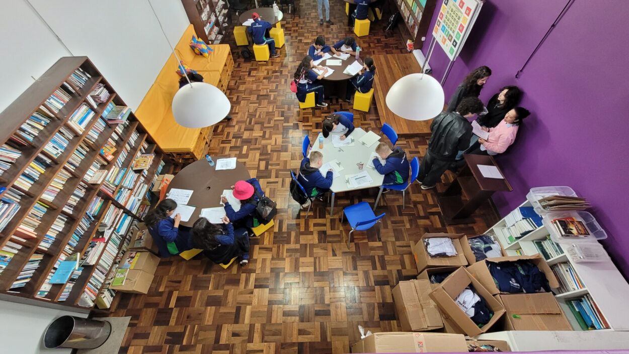 Vista superior da biblioteca da Escola Estadual Isolda Schmid. Estudantes estão distribuídos em mesas, realizando atividades de pesquisa, leitura, registro e discussão em grupo. O ambiente conta com estantes de livros ao longo das paredes e espaços destinados ao trabalho colaborativo.