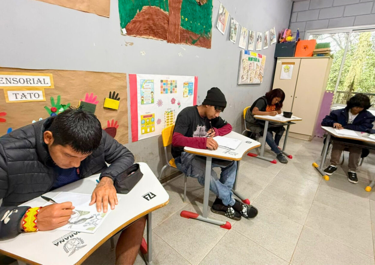 Quatro estudantes indígenas sentados em carteiras escolares dentro de uma sala de aula. Eles estão concentrados na produção de desenhos em folhas de papel apoiadas sobre as mesas. Em primeiro plano, um estudante desenha uma ilustração relacionada à pesquisa desenvolvida pelo grupo. Ao fundo, há cartazes pedagógicos fixados nas paredes, armários e uma janela com vista para a vegetação externa.