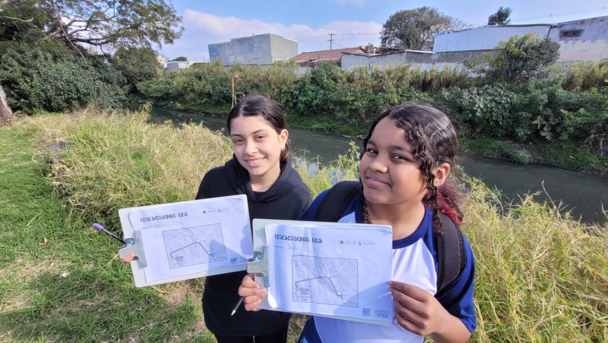 Duas estudantes seguram pranchetas com materiais de pesquisa durante atividade de campo às margens do Rio Belém, em Curitiba. Ao fundo, é possível ver o curso d'água cercado por vegetação e áreas urbanizadas. As alunas sorriem para a câmera enquanto registram informações relacionadas à investigação desenvolvida pelo Clube de Ciências Futuros Cientistas.