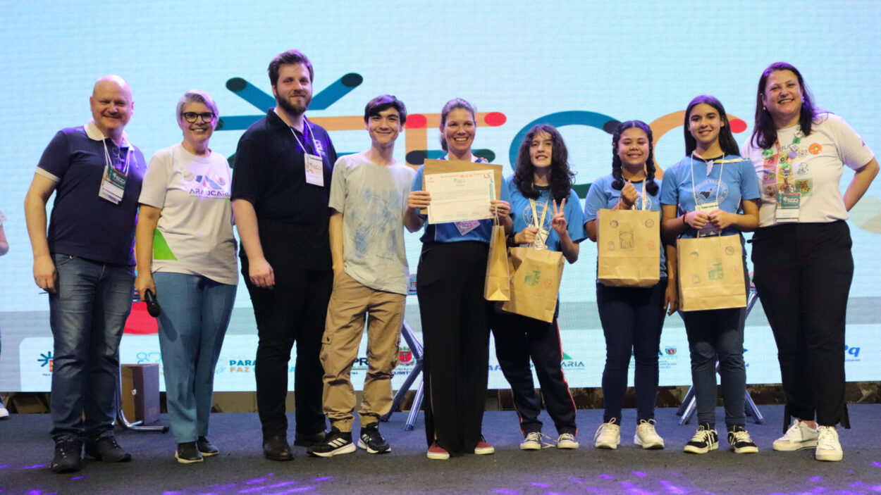 Foto mostra um grupo de estudantes e adultos no palco da FECCI. Ao centro, uma estudante segura um certificado, enquanto outros estudantes exibem medalhas e sacolas recebidas durante a premiação. Ao fundo, um painel colorido traz a identidade visual da feira.