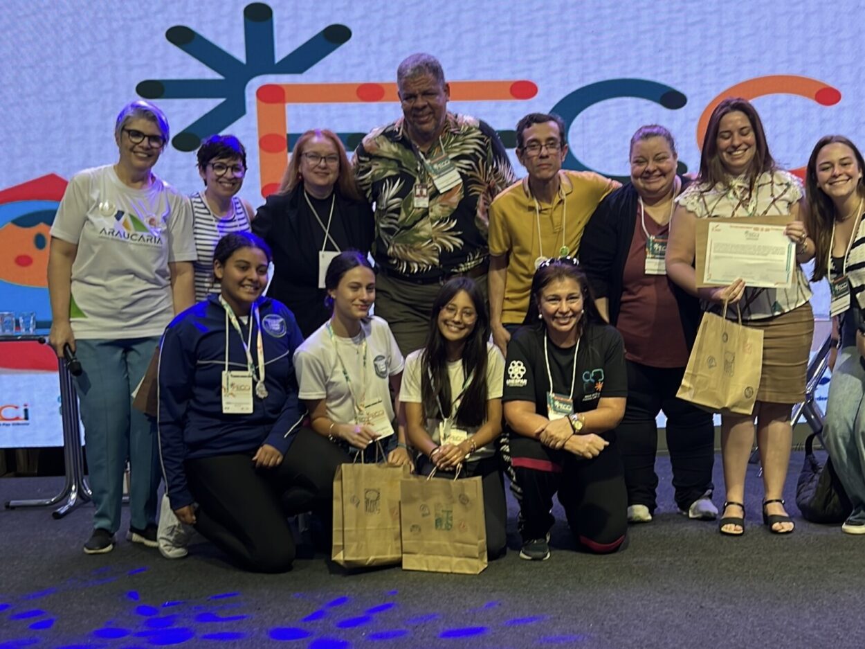 Foto mostra um grupo de estudantes e adultos no palco da FECCI. Ao fundo, um painel colorido traz a identidade visual da feira.