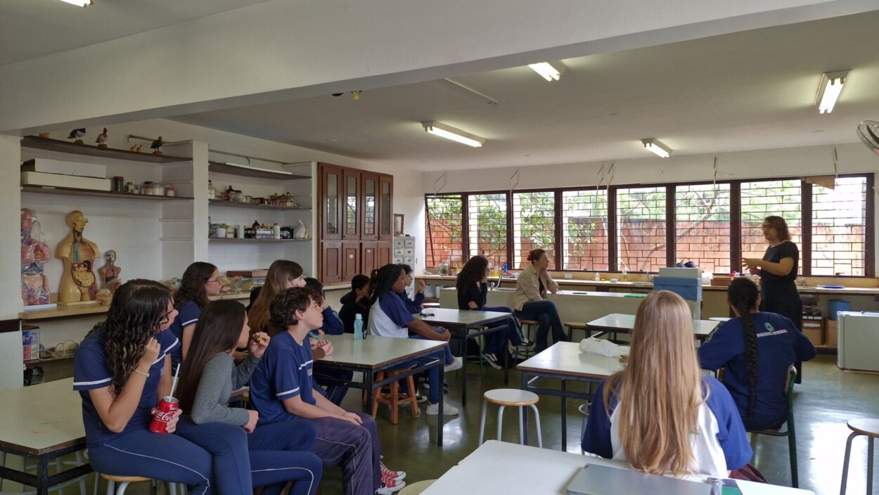 A imagem mostra um grupo de estudantes participando de uma atividade educativa em um laboratório de Ciências. Os alunos estão sentados ao redor de mesas, atentos à explicação de uma professora que conduz a atividade na parte frontal da sala. O ambiente conta com bancadas, armários e diversos materiais didáticos, incluindo modelos anatômicos do corpo humano e outros recursos utilizados para o ensino científico.