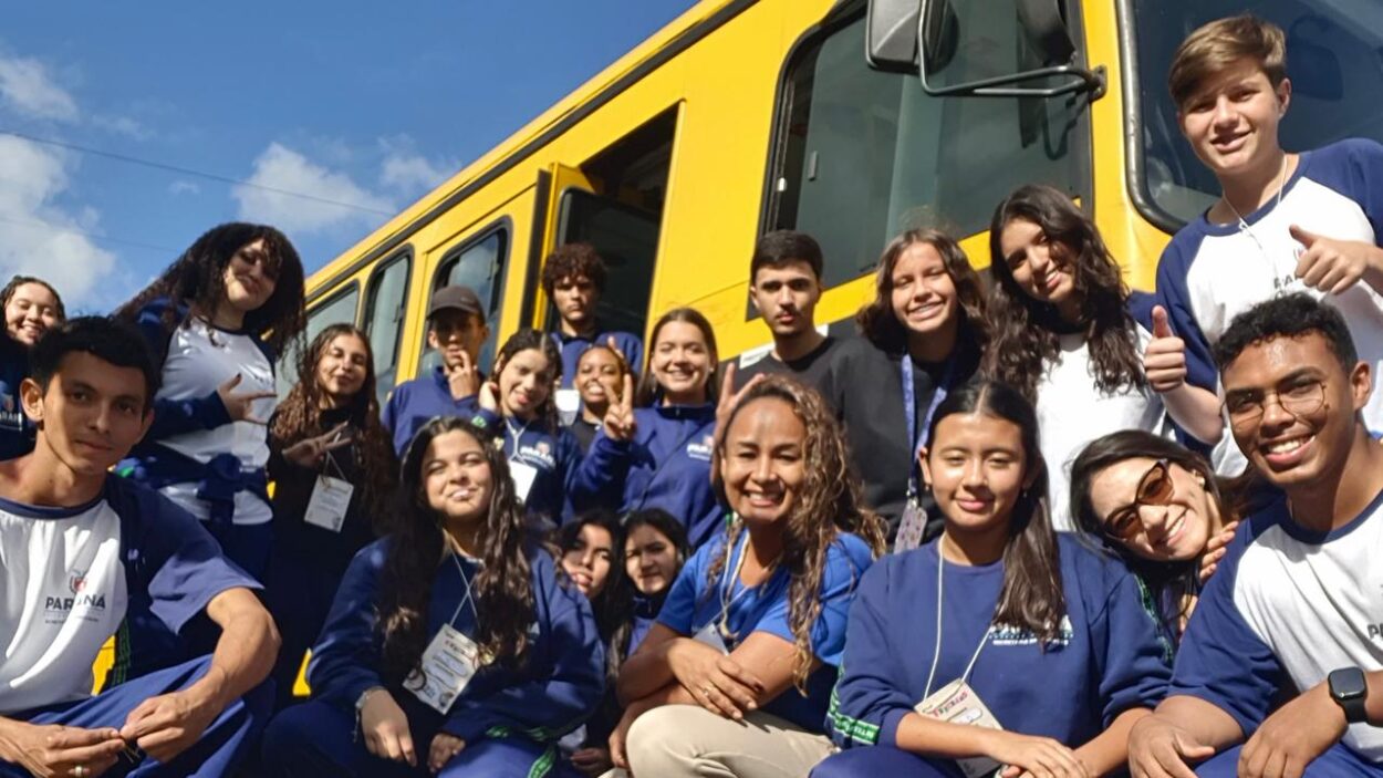 Foto em grupo ao ar livre, em frente a um ônibus amarelo escolar. Cerca de vinte adolescentes e duas professoras posam juntos sorrindo para a câmera. A maioria usa uniforme azul e branco da escola, alguns com crachá pendurado no pescoço. Os alunos estão distribuídos em diferentes alturas: alguns agachados na frente, outros em pé ao lado e próximos à porta do ônibus. Vários fazem sinais com as mãos ou mostram joinha. O céu azul aparece ao fundo, com poucas nuvens, e a luz do sol ilumina a cena, criando um clima alegre e descontraído de passeio ou excursão escolar.