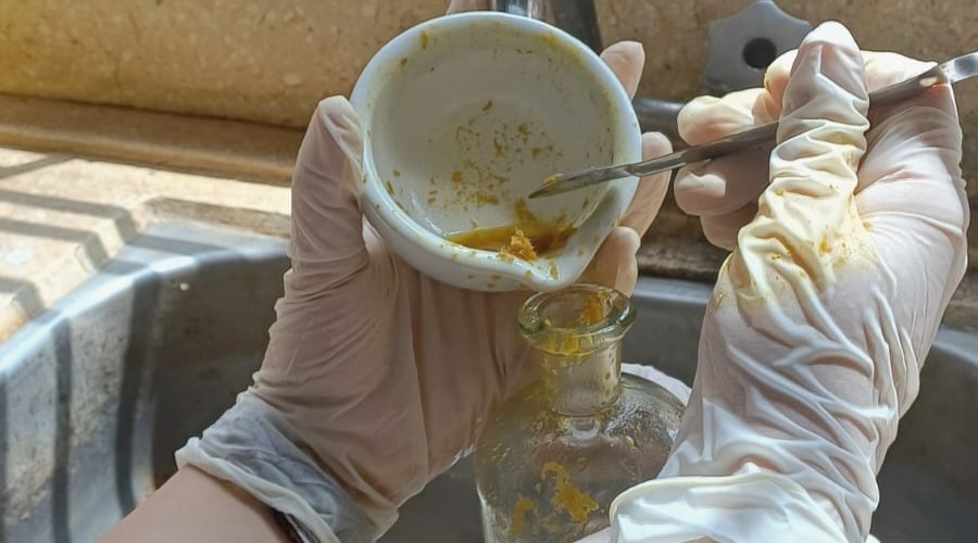 A imagem mostra uma atividade experimental em ambiente laboratorial. Uma pessoa utilizando luvas de proteção manipula uma substância amarelada com auxílio de uma espátula e um almofariz de porcelana, transferindo o material para um recipiente de vidro. A cena evidencia cuidados com segurança e procedimentos de laboratório, demonstrando uma etapa de preparação, análise ou processamento de amostras.