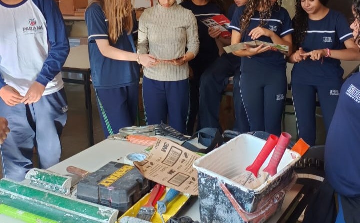 A imagem mostra um grupo de estudantes reunidos em uma sala de aula ou laboratório, participando de uma atividade prática. No centro da cena, há uma mesa com diversos materiais e ferramentas, como bandejas, pincéis, espátulas, lixas, caixas organizadoras e outros equipamentos utilizados em oficinas ou experimentos. Os alunos observam atentamente os objetos enquanto seguram materiais de leitura, sugerindo um momento de aprendizagem investigativa e discussão sobre técnicas, procedimentos ou aplicações científicas.