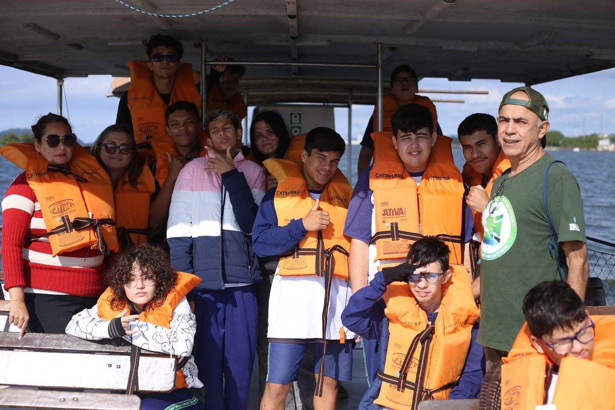 Fotografia colorida feita durante um passeio de barco na Baía de Antonina. Em primeiro plano, um grupo de estudantes e educadores está reunido na embarcação usando coletes salva-vidas laranja. Os participantes posam para a câmera em diferentes expressões, enquanto ao fundo é possível ver a estrutura coberta do barco e o horizonte da baía sob a luz do dia. A atividade integra ações de observação científica dos manguezais e da biodiversidade da região.