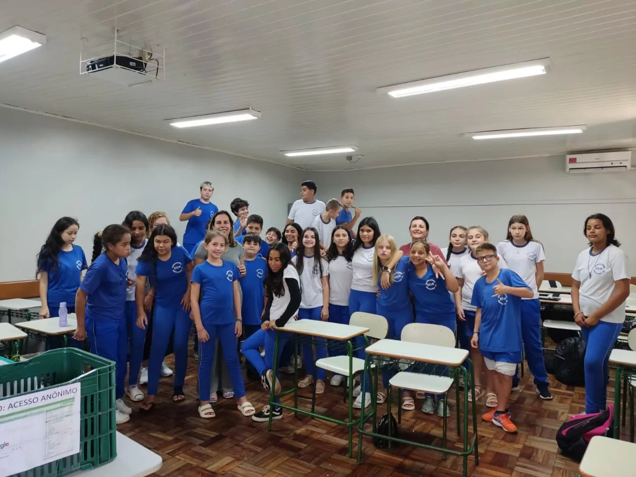 A foto mostra o grupo de estudantes em uma sala de aula. A maioria dos alunos veste uniformes escolares, alguns usando roupas azuis e outros, brancas. Eles estão juntos para uma foto em grupo, com sorrisos e poses descontraídas. O ambiente é uma sala de aula comum, com cadeiras e mesas organizadas, e há um ar condicionado no canto superior direito. O teto possui luminárias retangulares, e na parte superior esquerda há um projetor suspenso. O chão é de madeira.
