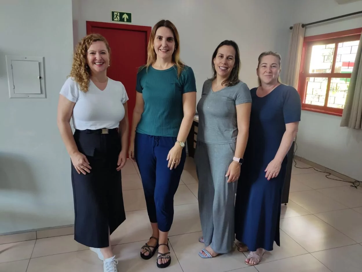 A foto mostra quatro pessoas posando para uma foto em um ambiente interno bem iluminado. Todos estão sorrindo e de pé lado a lado. A pessoa à esquerda tem cabelos cacheados loiros e está vestindo uma camiseta branca e saia preta com tênis branco. A segunda pessoa, da esquerda para a direita, tem cabelos lisos e usa uma camiseta verde escura e calça azul marinho, com sandálias. A terceira pessoa veste um conjunto cinza, com blusa e calça largas, e usa um relógio no pulso esquerdo. A pessoa à direita tem cabelos presos, veste um vestido azul escuro e sandálias claras. Ao fundo, há uma porta vermelha e uma janela, além de uma placa de saída de emergência verde acima da porta. O ambiente parece ser um espaço formal, com piso claro e paredes brancas.