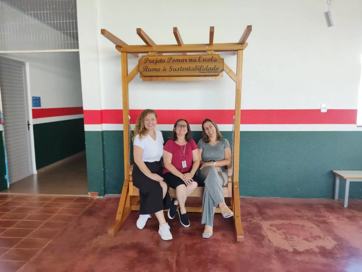 A foto mostra três pessoas sentadas em um banco de madeira suspenso por correntes, estilo balanço, sob uma estrutura de madeira. Acima delas, há uma placa que diz "Projeto Pomar na Escola Rumo à Sustentabilidade" com o período letivo 2023-2024. A pessoa à esquerda tem cabelos cacheados loiros, veste uma camiseta branca, saia preta e tênis branco. A pessoa no centro usa óculos, uma camiseta vermelha e calça preta, e está sorrindo. A pessoa à direita veste um conjunto cinza de blusa e calça e também está sorrindo. É um ambiente escolar, com paredes pintadas em verde, vermelho e branco, e o chão de cimento ou tijolos rústicos. Ao fundo, há uma entrada com uma porta aberta.