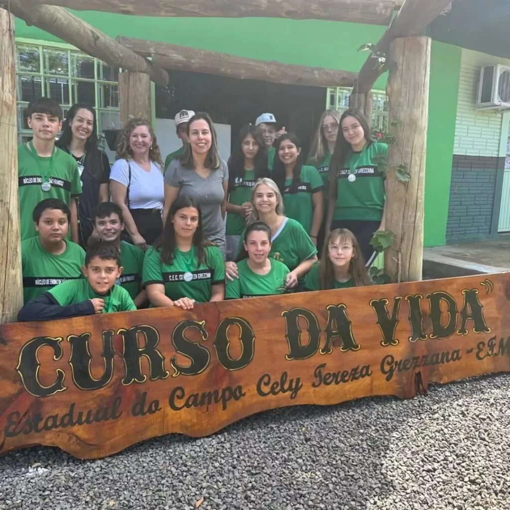 A foto mostra o grupo de pessoas reunidas em frente a uma estrutura de madeira com uma placa grande que contém os dizeres "CURSO DA VIDA" e "Estadual do Campo Cely Tereza Grezzana - EEM". A maioria das pessoas está vestindo camisetas verdes com uma faixa preta contendo texto. Algumas pessoas estão em pé, enquanto outras estão agachadas ou sentadas à frente da placa. O ambiente parece ser uma área externa, com um prédio ao fundo pintado de verde e branco, e há vegetação ao redor. Todos estão posando para a foto, demonstrando expressões amigáveis.