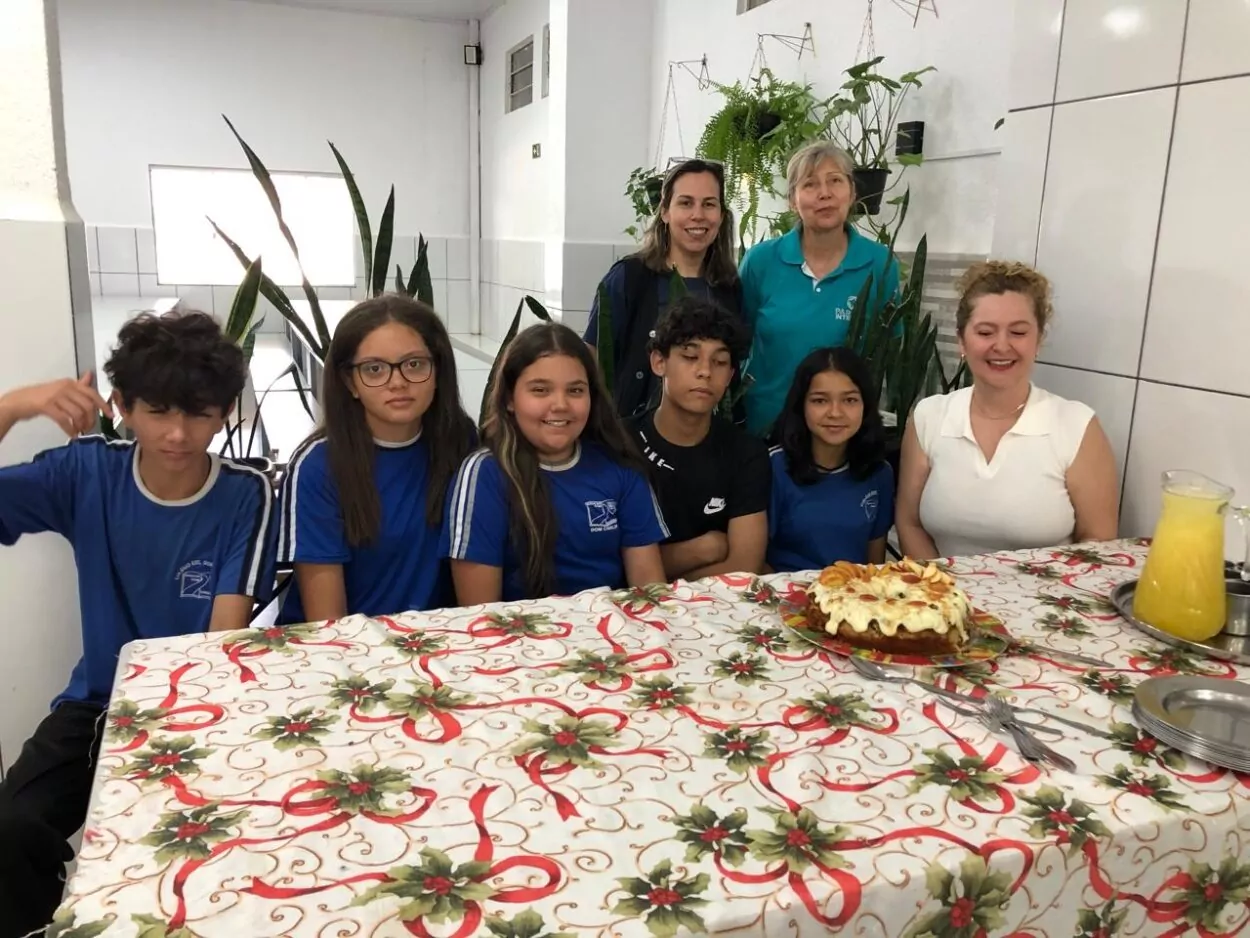 A foto mostra um grupo de pessoas reunidas ao redor de uma mesa coberta com uma toalha decorada com temas natalinos. Sobre a mesa, há um bolo decorado com frutas e uma jarra de suco amarelo. Algumas pessoas estão sentadas, enquanto outras estão de pé atrás delas. Algumas vestem uniformes escolares com camisetas azuis com detalhes brancos, enquanto outras usam roupas casuais. O ambiente é um espaço fechado, com paredes brancas, azulejos e plantas penduradas e também no chão. As expressões variam entre sorrisos e neutralidade.