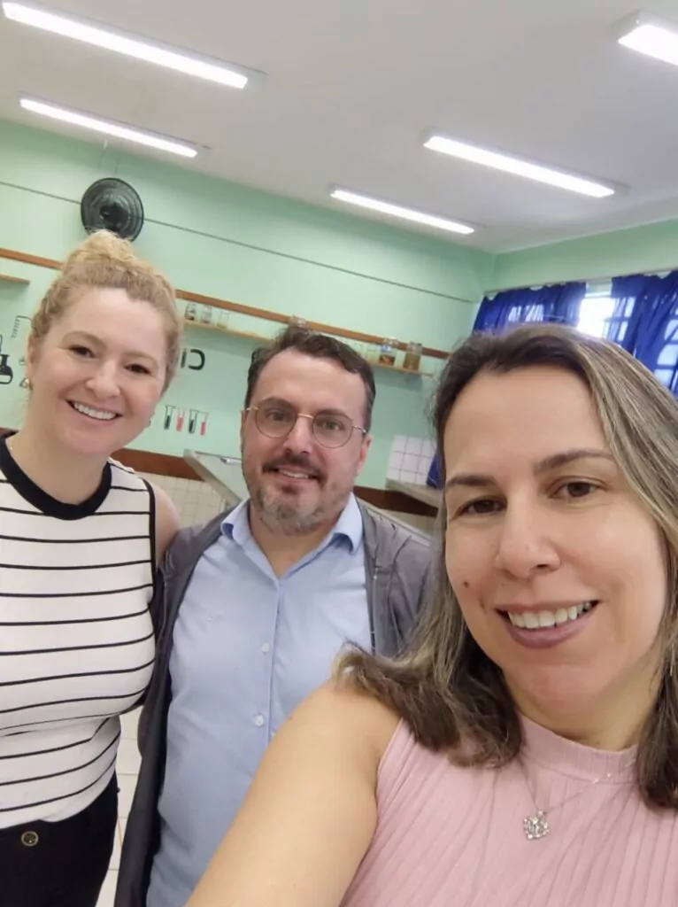 A foto mostra três pessoas sorrindo para uma selfie tirada em um ambiente interno. O local parece ser uma sala de laboratório ou de ensino, com paredes verdes, prateleiras com frascos e materiais, além de bancadas e utensílios pendurados na parede. Há uma cortina azul cobrindo parcialmente as janelas, e luminárias no teto iluminam o ambiente. As pessoas na foto vestem roupas casuais e sociais, demonstrando expressões amigáveis e descontraídas.