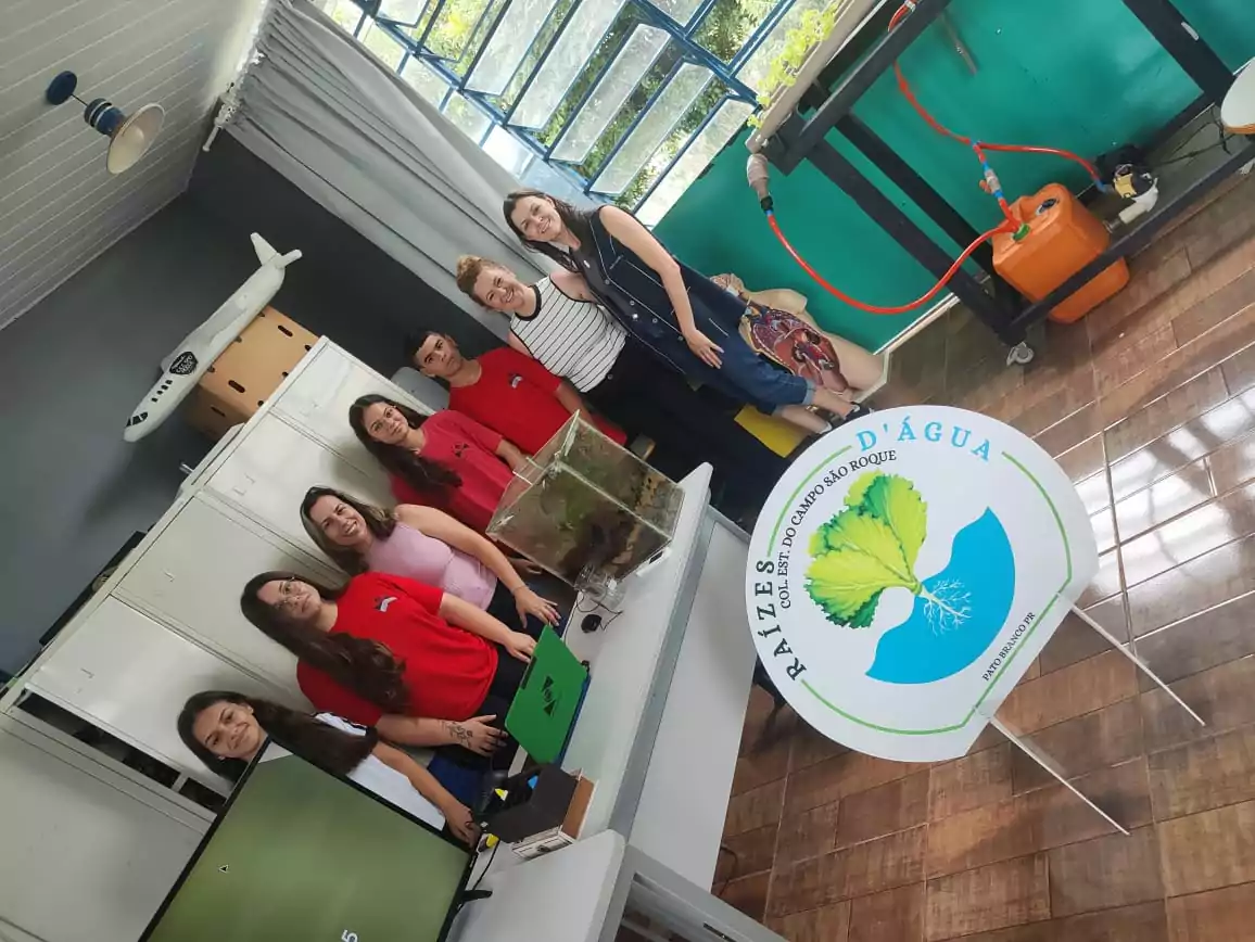 A foto mostra um grupo de pessoas posando para uma foto em um ambiente que parece ser uma sala de aula ou laboratório escolar. Algumas pessoas estão vestindo camisetas vermelhas com um pequeno logotipo, enquanto outras usam roupas casuais. No primeiro plano, há um painel circular com o nome "Raízes D'Água" e a inscrição "Col. Est. do Campo São Roque". O ambiente contém armários brancos, computadores e um terrário sobre uma mesa. No fundo, há uma bancada com tubulações e equipamentos, além de um manequim vestido com roupas típicas. Também é possível ver um modelo de avião branco fixado na parede, e janelas grandes permitem a entrada de luz natural. O grupo está sorrindo.