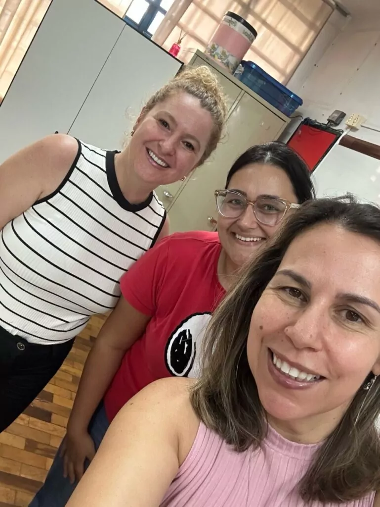 A foto mostra três pessoas sorrindo para uma selfie em um ambiente interno, possivelmente uma sala de aula ou escritório. No fundo, há armários metálicos, caixas plásticas e um recipiente grande sobre um dos móveis. O piso é de madeira e as cortinas estão fechadas, filtrando a luz que entra pela janela. As pessoas na foto demonstram expressões alegres e estão vestindo roupas casuais. Uma delas veste uma blusa listrada, outra usa uma camiseta vermelha com um símbolo circular, e a terceira usa uma blusa rosa.