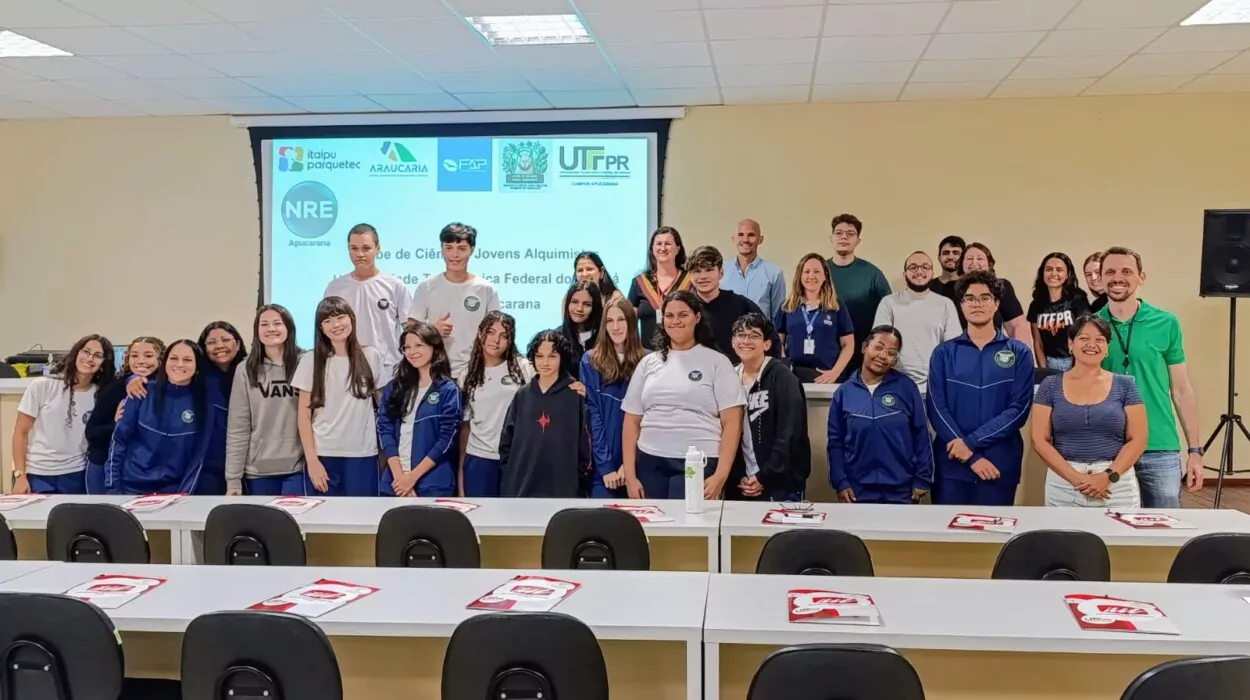 A foto mostra um grupo grande de pessoas reunidas em um auditório. Ao fundo, há um telão com projeção que indica o evento: trata-se de uma atividade promovida por diversas instituições, incluindo a UTFPR (Campus Apucarana), Itaipu Parquetec, Fundação Araucária, FAP e o NRE Apucarana, com o título "Clube de Ciências Jovens Alquimistas". Estão presentes estudantes do clube de ciências e professores. Os estudantes usam uniformes escolares ou roupas casuais, e muitos deles estão sorrindo, mostrando um clima descontraído.Nas mesas em primeiro plano, há cadernos personalizados. O ambiente é bem iluminado, com teto de placas e luzes fluorescentes. A foto transmite uma atmosfera de integração entre escola e universidade, com foco em educação científica e incentivo ao conhecimento.