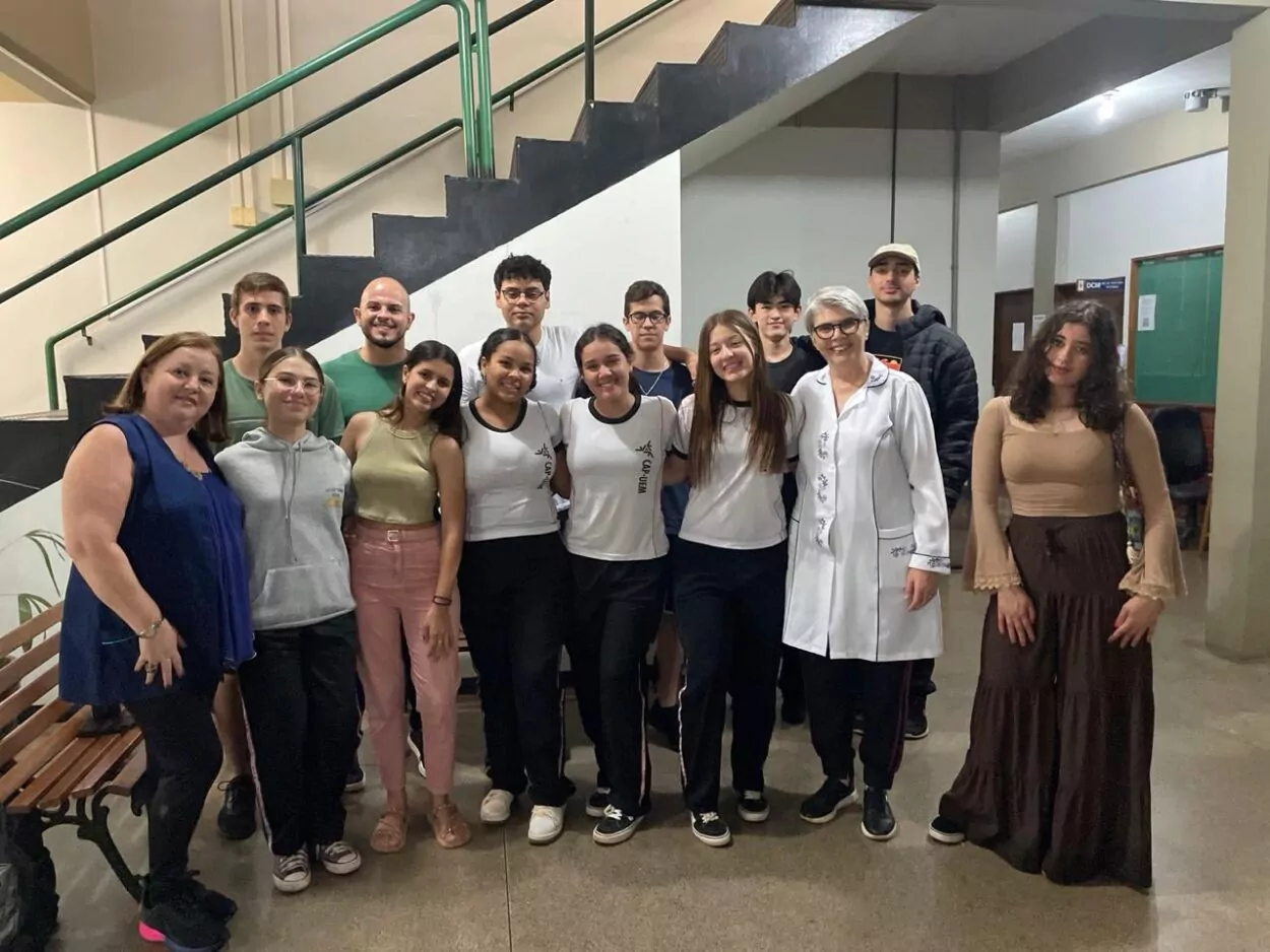 A imagem mostra um grupo de pessoas posando para uma foto em um ambiente escolar, próximo a uma escada. O grupo é composto por alunos e os professores: Antonio de Oliveira, Rosemary Margarida Campos Parizotto e Débora Sant’Ana, todos sorrindo para a câmera. Alguns alunos estão usando uniformes escolares brancos com detalhes pretos, enquanto outros estão vestidos de forma casual. A atmosfera parece amigável e descontraída. O ambiente conta com bancos, paredes claras e um corredor ao fundo.