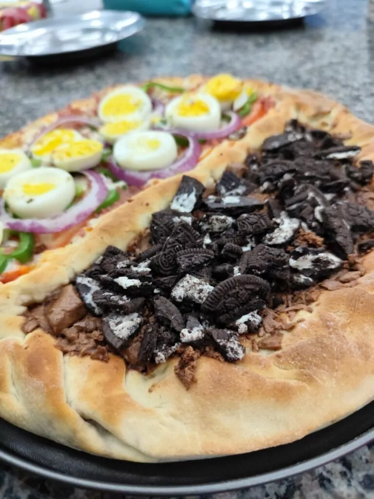 A imagem mostra uma pizza de dois sabores servida sobre um prato preto em uma superfície de granito. A metade esquerda tem cobertura salgada, incluindo ovos cozidos fatiados, rodelas de cebola roxa, pimentão e queijo derretido. Já a metade direita é doce, coberta com pedaços de chocolate derretido e biscoitos tipo Oreo quebrados. No fundo, há pratos metálicos sobre a mesa, sugerindo um ambiente de cozinha ou laboratório culinário