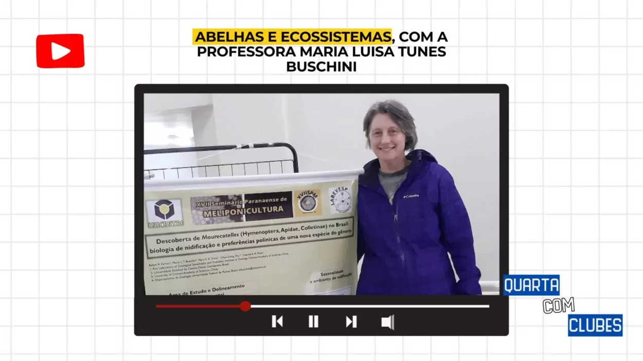 Fotografia de uma sala de aula com janelas grandes ao fundo, por onde entra luz natural. Um grupo de sete pessoas está reunido, sentadas em carteiras escolares organizadas em semicírculo. À esquerda, um homem de barba, usando camiseta azul, está sentado e gesticulando enquanto fala. À sua frente, os outros participantes — três mulheres e três homens — prestam atenção, alguns com cadernos ou notebooks no colo. Há duas garrafas de refrigerante e alguns lanches sobre uma cadeira próxima à parede. A sala é clara, com piso branco e cadeiras azuis. Ao fundo, é possível ver o pátio externo e alguns prédios.
