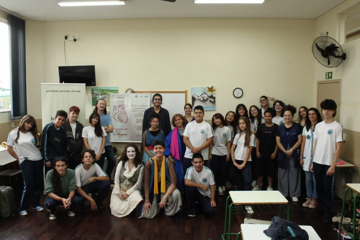 A imagem mostra um grupo numeroso de pessoas reunidas dentro de uma sala de aula para uma foto coletiva. A maioria veste uniformes escolares brancos com detalhes verdes ou azulados, enquanto algumas pessoas usam roupas de cena ou figurinos teatrais, incluindo maquiagem artística. Ao fundo, há um quadro branco com cartazes afixados, um deles com o desenho de um coração anatômico e os dizeres “Coração em Chagas”. Também há pinturas decorativas na parede, um relógio redondo, uma televisão e ventiladores. A atmosfera é descontraída e alegre, sugerindo um encontro entre estudantes, docentes e artistas após uma apresentação ou atividade cultural ligada à ciência e arte.