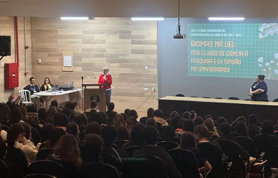 A imagem mostra um auditório com estudantes assistindo à palestra de abertura do evento realizado na UEL. Uma mulher com blusa vermelha está em pé falando ao microfone. À esquerda, duas pessoas estão sentadas acompanhando a apresentação. No canto direito do palco, uma mulher está observando a fala. No telão, está projetado o título do evento: "Biomas na UEL: dos clubes de ciência à pesquisa e extensão na universidade", com o subtítulo: "UEM e a rede paranaense de popularização da ciência na SNCT 2024". O ambiente é fechado, com iluminação artificial, paredes revestidas com paineis de madeira clara e cadeiras pretas.