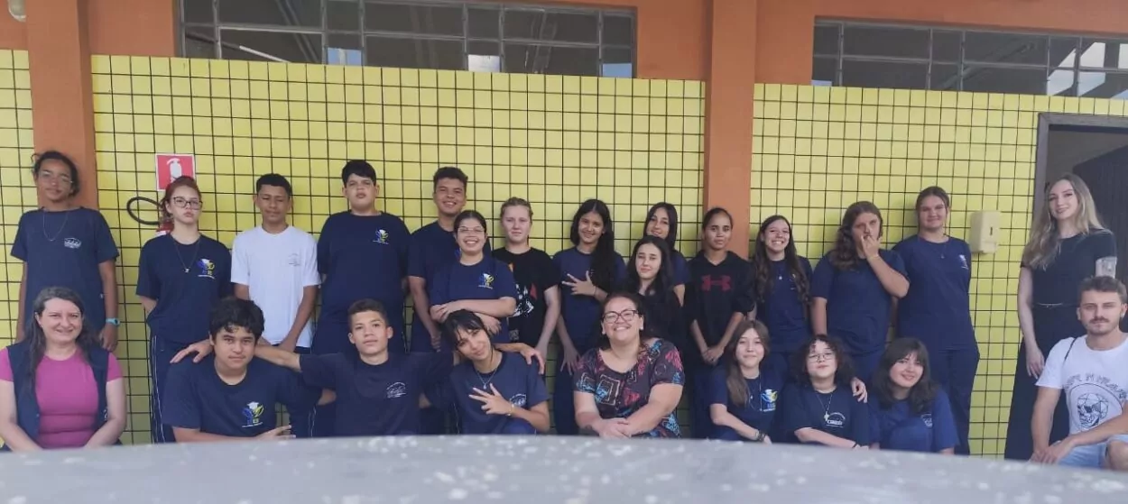 Essa imagem mostra um grupo de estudantes do clube de ciência "Nutrindo Mentes", da Escola Estadual Espírito Santo, em Ponta Grossa. Estão organizados em duas fileiras — a da frente sentada e a de trás em pé — posando para a foto com expressões descontraídas e sorridentes. Atrás deles, há uma parede com revestimento amarelo em pastilhas, típica de prédios escolares. Ao lado direito da imagem, aparecem duas pessoas adultas, professores ou orientadores do clube.