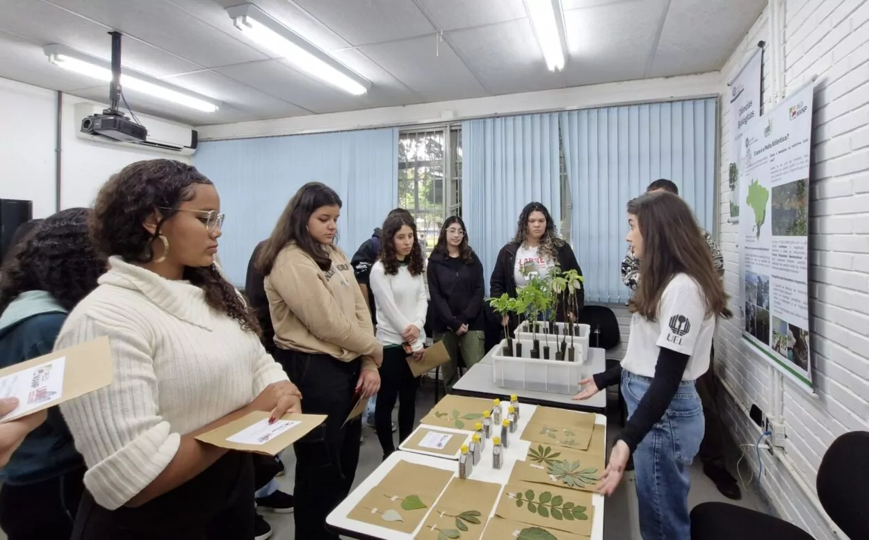 A imagem mostra um grupo de estudantes em uma sala, visitando um estande sobre biologia vegetal. Todos estão de pé, em semicírculo ao redor de uma mesa retangular coberta com materiais didáticos. Na mesa, há várias folhas secas organizadas e coladas em folhas de papel kraft, frascos pequenos com líquidos e alguns materiais informativos. Também há uma caixa plástica contendo mudas de plantas. À direita da imagem, uma mulher jovem com cabelos castanhos, vestindo uma camiseta branca com o logo da UEL (Universidade Estadual de Londrina) e calça jeans, está explicando o conteúdo para os alunos. Ao fundo, há cartazes na parede com informações sobre biodiversidade vegetal no Brasil, mapas e imagens de áreas naturais.