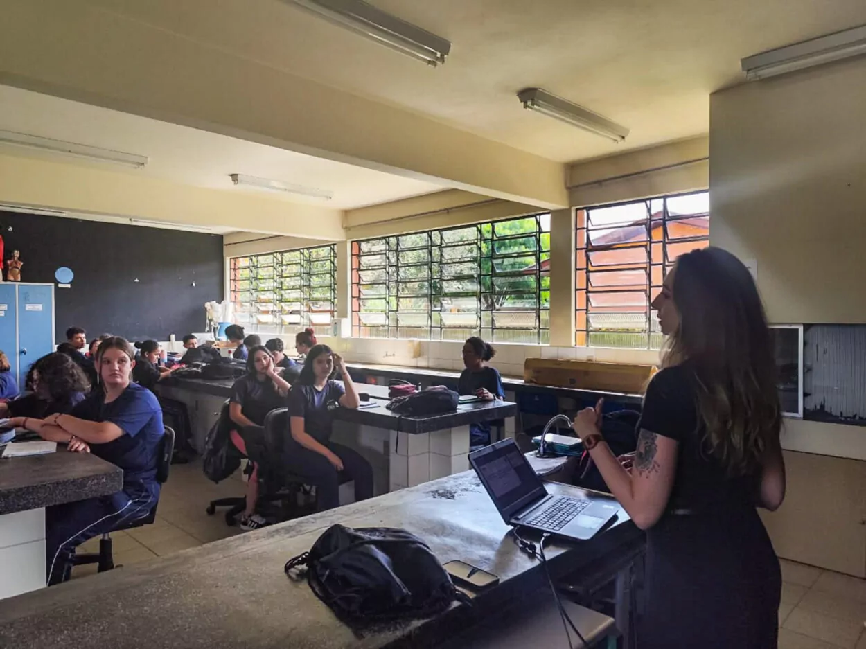 A foto mostra uma sala de aula com vários estudantes sentados em bancadas que parecem ser de um laboratório. Eles estão prestando atenção na pessoa em pé à frente da sala, que está realizando uma apresentação utilizando um laptop. A iluminação natural entra pelas janelas amplas ao fundo, e há armários ao lado esquerdo da sala. A atmosfera parece ser de concentração, com os alunos voltados para a apresentação.
