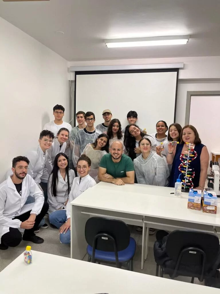 A imagem mostra um grupo de pessoas reunidas no Laboratório de Imunogenética. Entre eles, há estudantes, professores e profissionais vestidos com jalecos brancos. Todos estão sorrindo e posando para a foto, com um homem de camiseta verde ao centro apoiado em uma mesa. No ambiente, há uma tela de projeção ao fundo, microscópio, caixas de leite e um modelo de DNA colorido sobre a mesa, indicando atividades ligadas à ciência e biologia. A atmosfera parece descontraída e colaborativa, sugerindo um evento educacional.